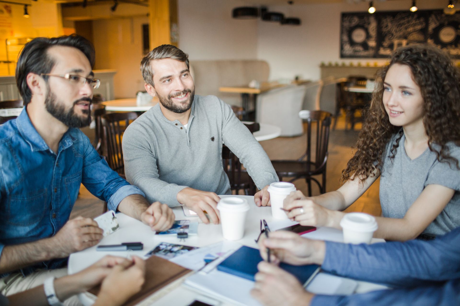 Four people sit around a table in a cafe, talking and holding coffee cups.