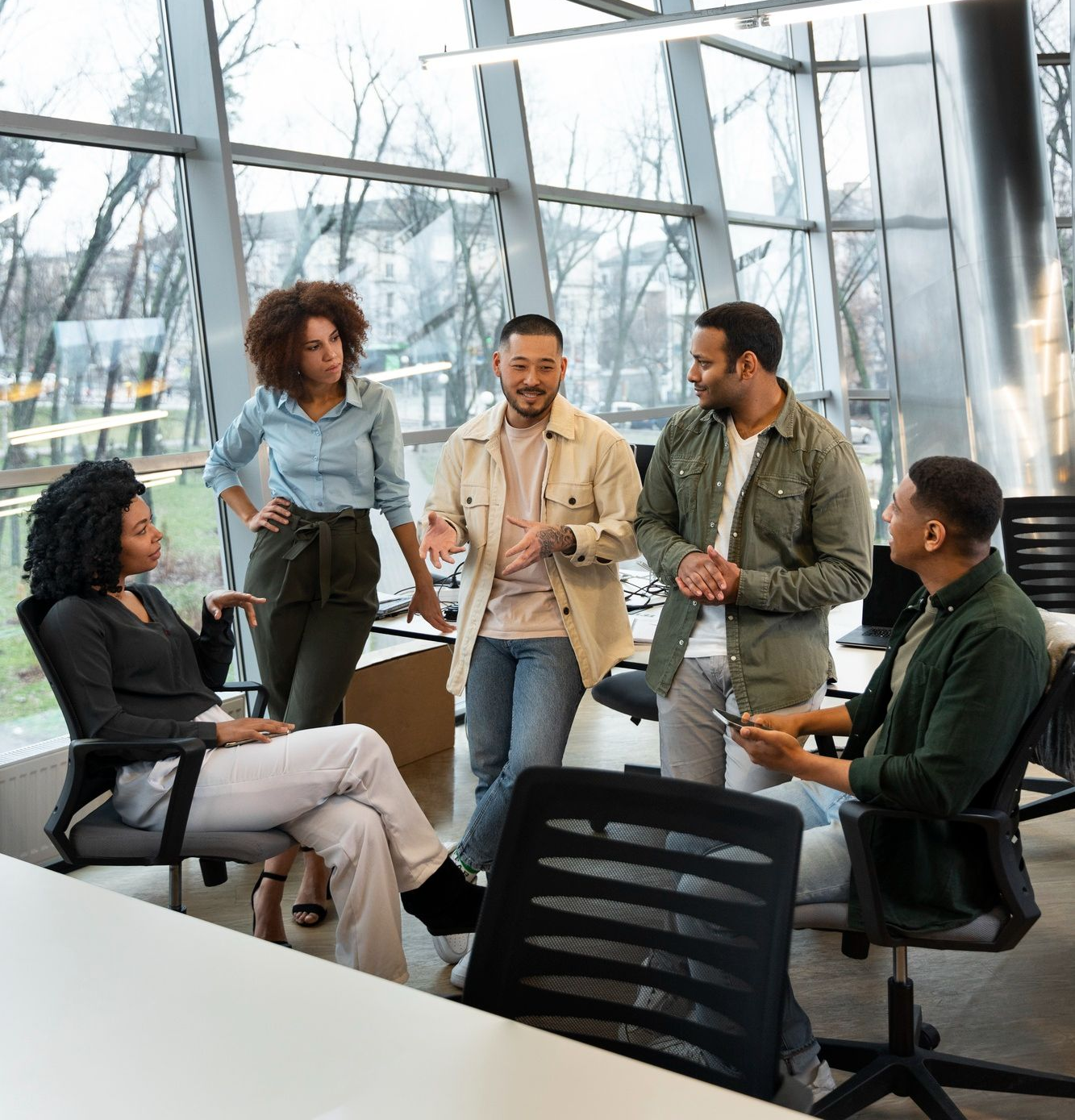 Group of people in an office, engaged in a discussion near large windows.