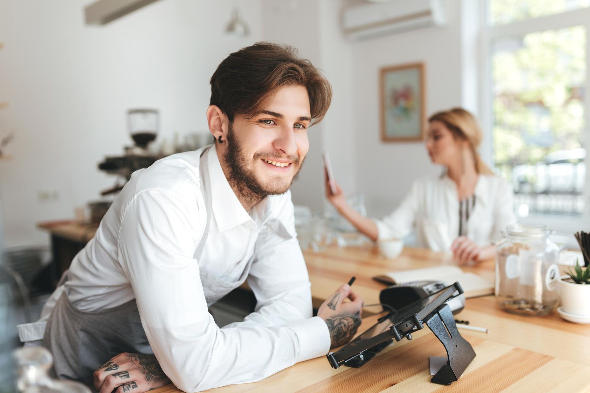 Man smiling, leaning on a counter, with a card reader. Woman in the background. Coffee shop setting.