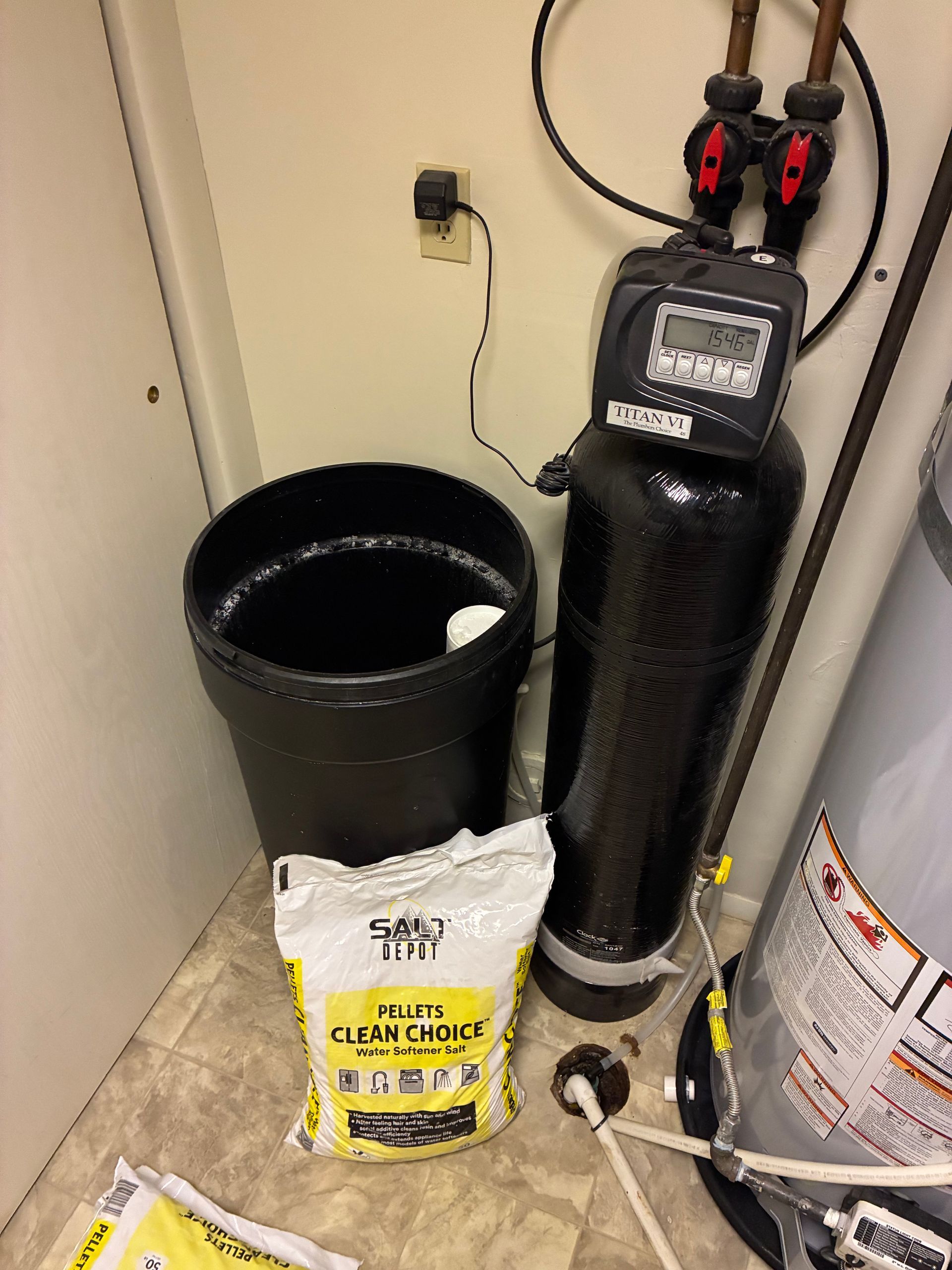 Water softener system beside a black trash can and salt bag in a utility room.