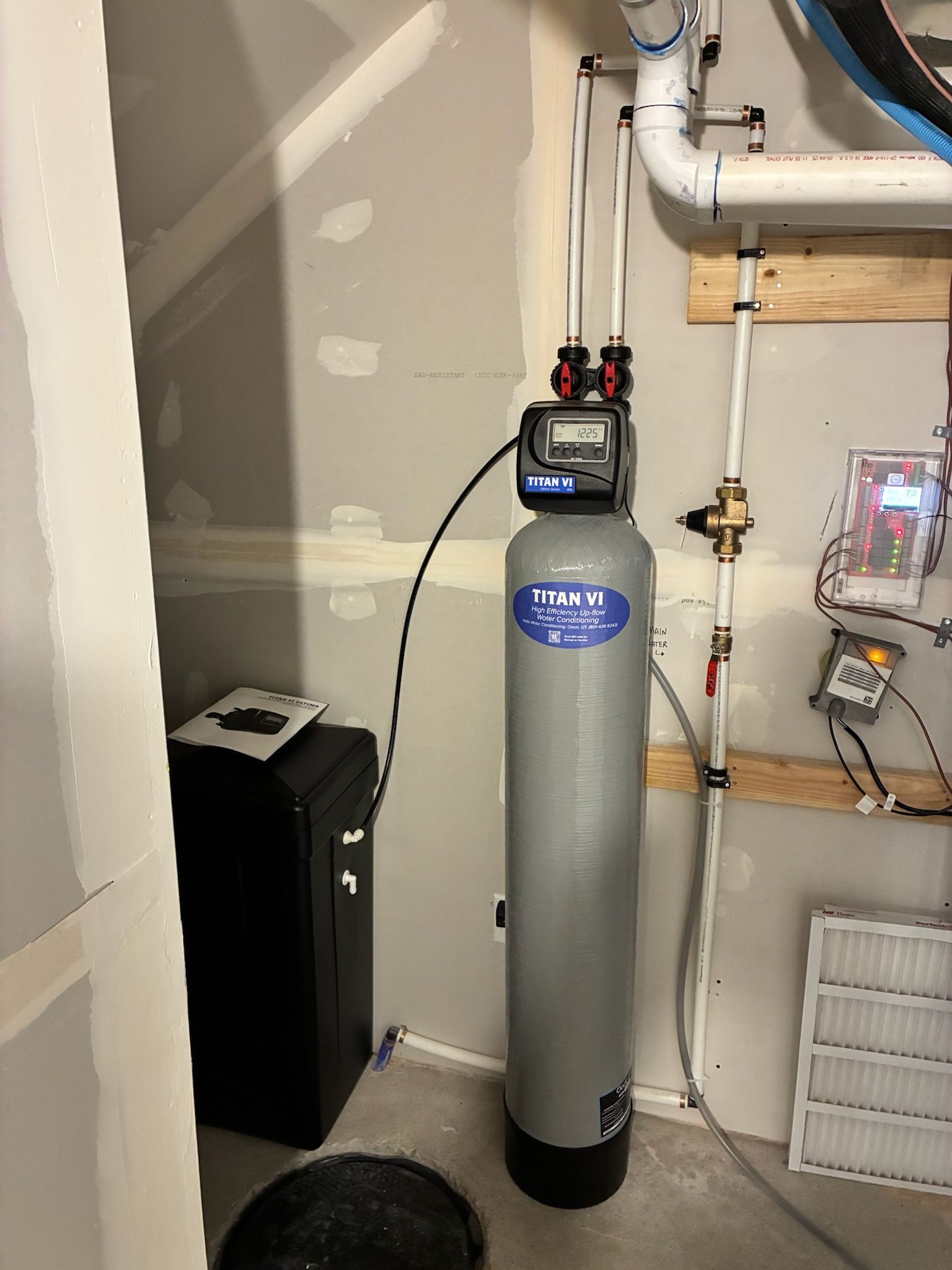 Water softener tank and utility equipment in a basement corner with exposed pipes and cables.