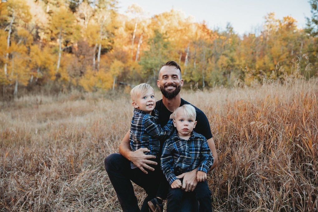 Man kneels in a grassy field with two young children, autumn trees glowing in the background.