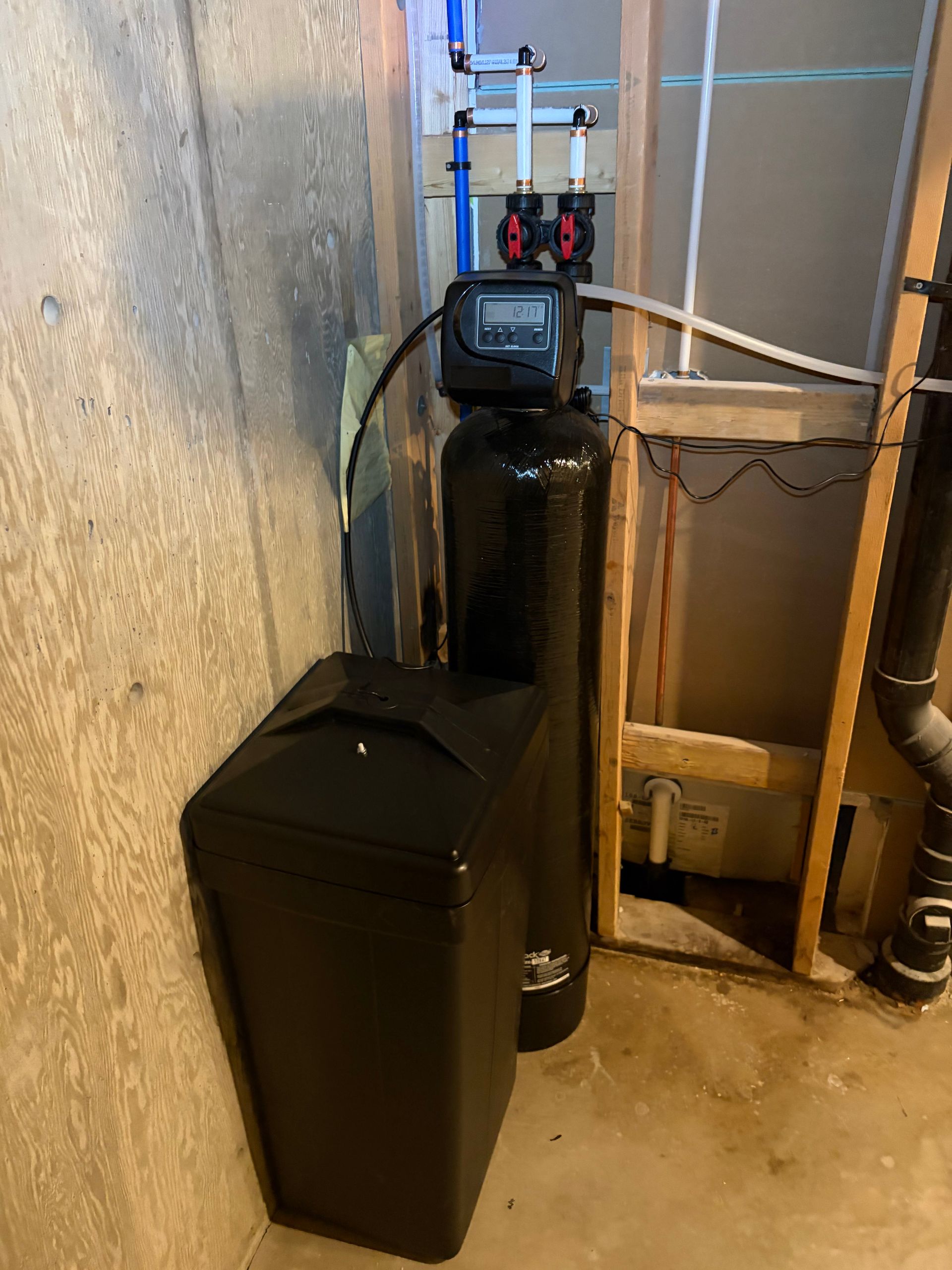 Utility room with a black water softener tank and cabinet beside exposed pipes and valves.
