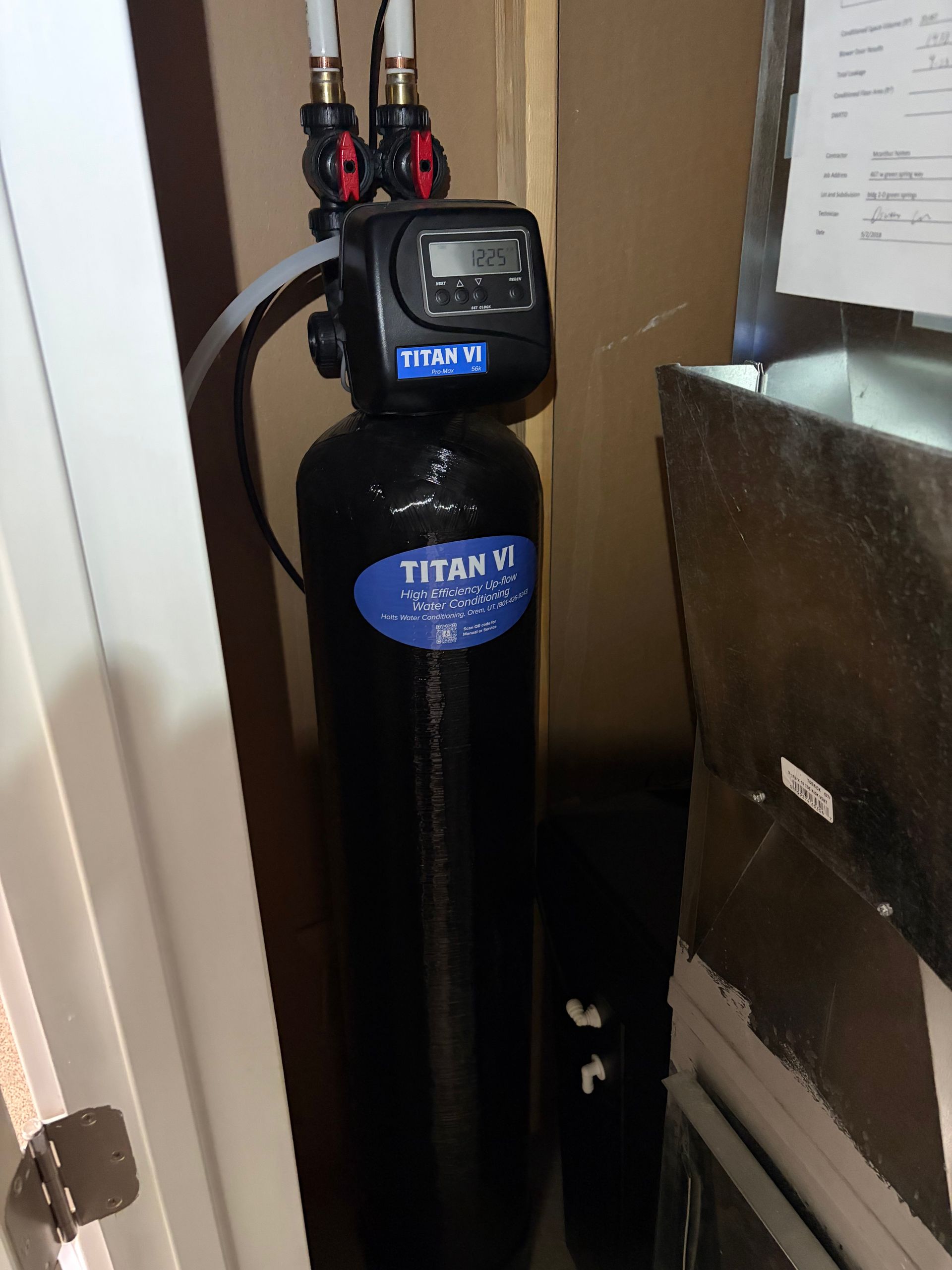Black water softener tank with digital control head in a utility room, beside cardboard boxes.
