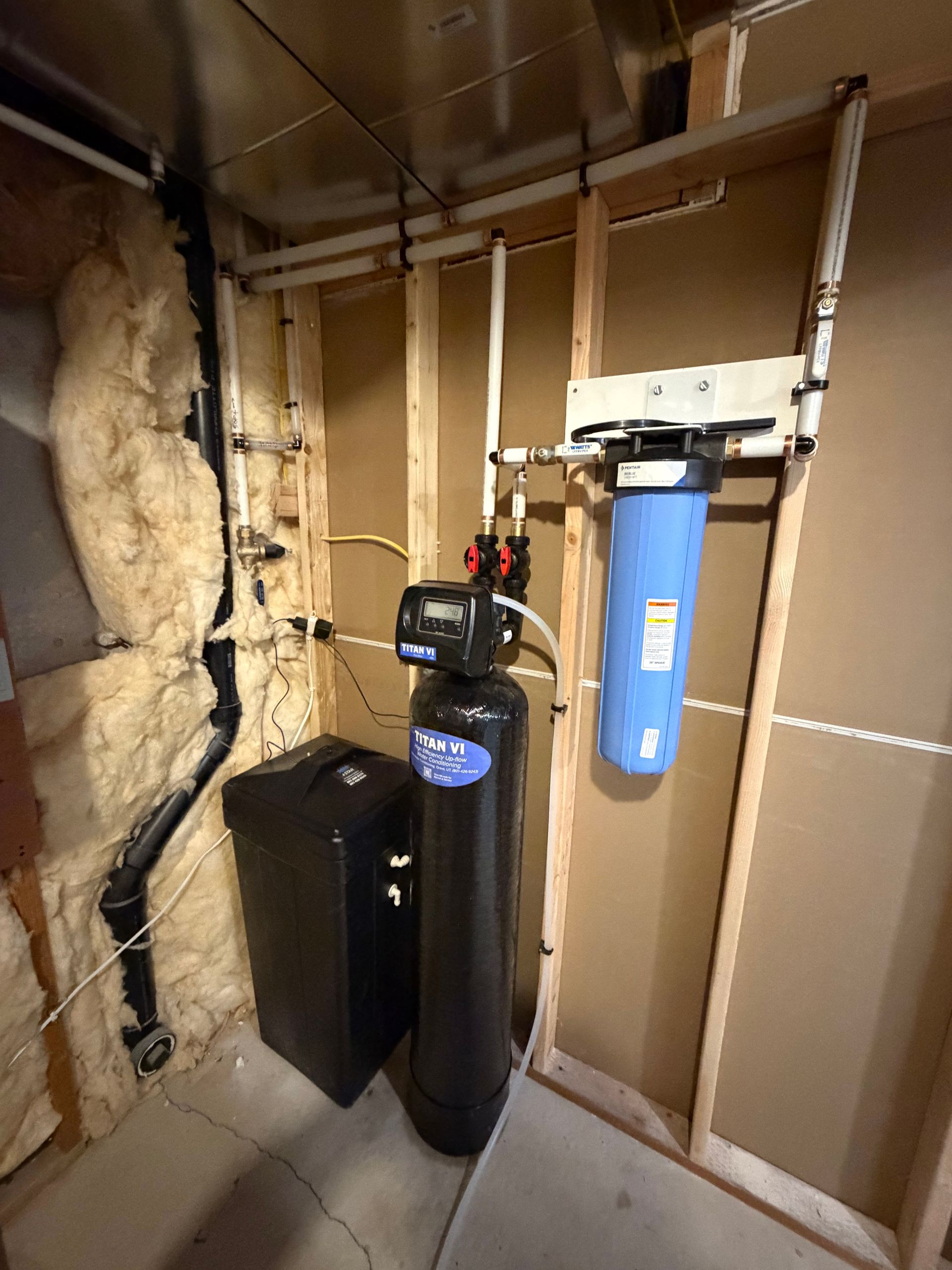Basement utility area with water softener, blue filter tank, pipes, and exposed insulation on unfinished walls
