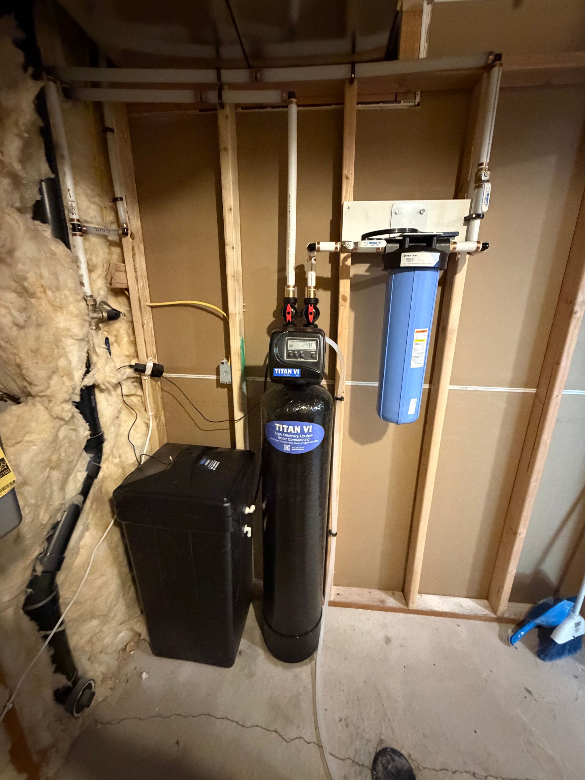 Water softener and filtration tank in an unfinished basement beside exposed wall insulation and framing