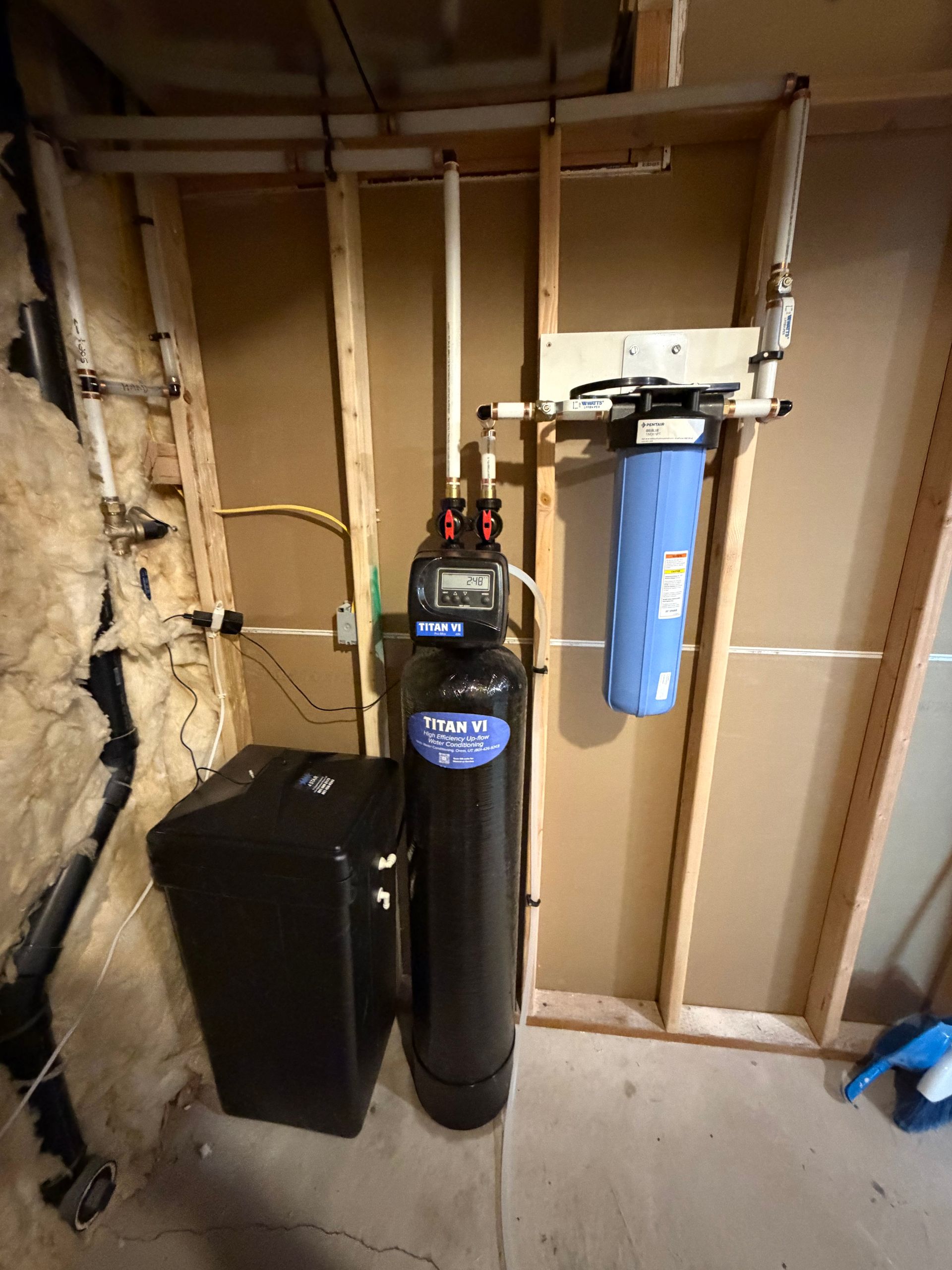 Basement utility area with a water softener, black tank, and blue filter mounted on exposed wall framing.