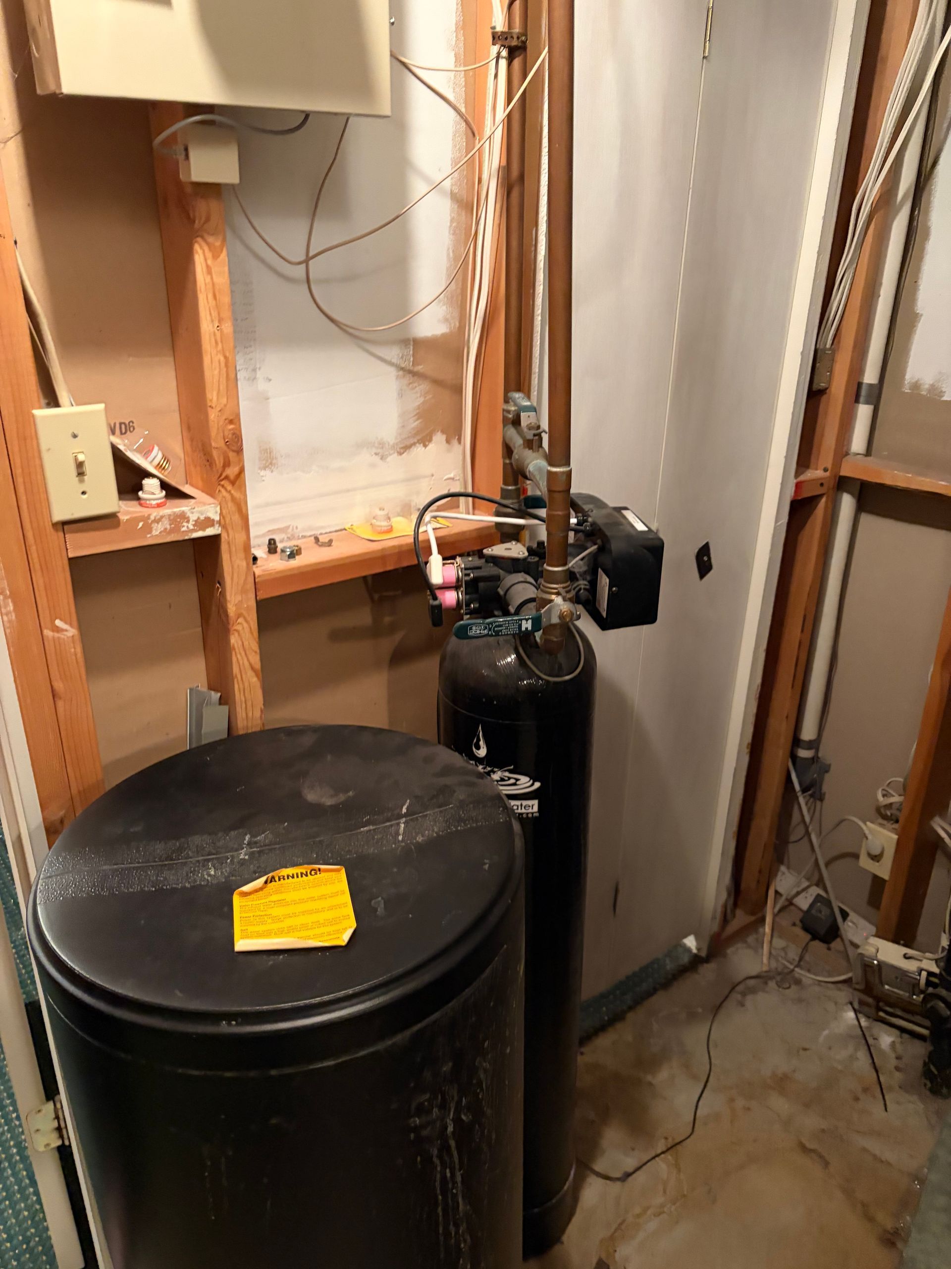 Water heater and black storage tank in a utility closet with exposed pipes and wiring.