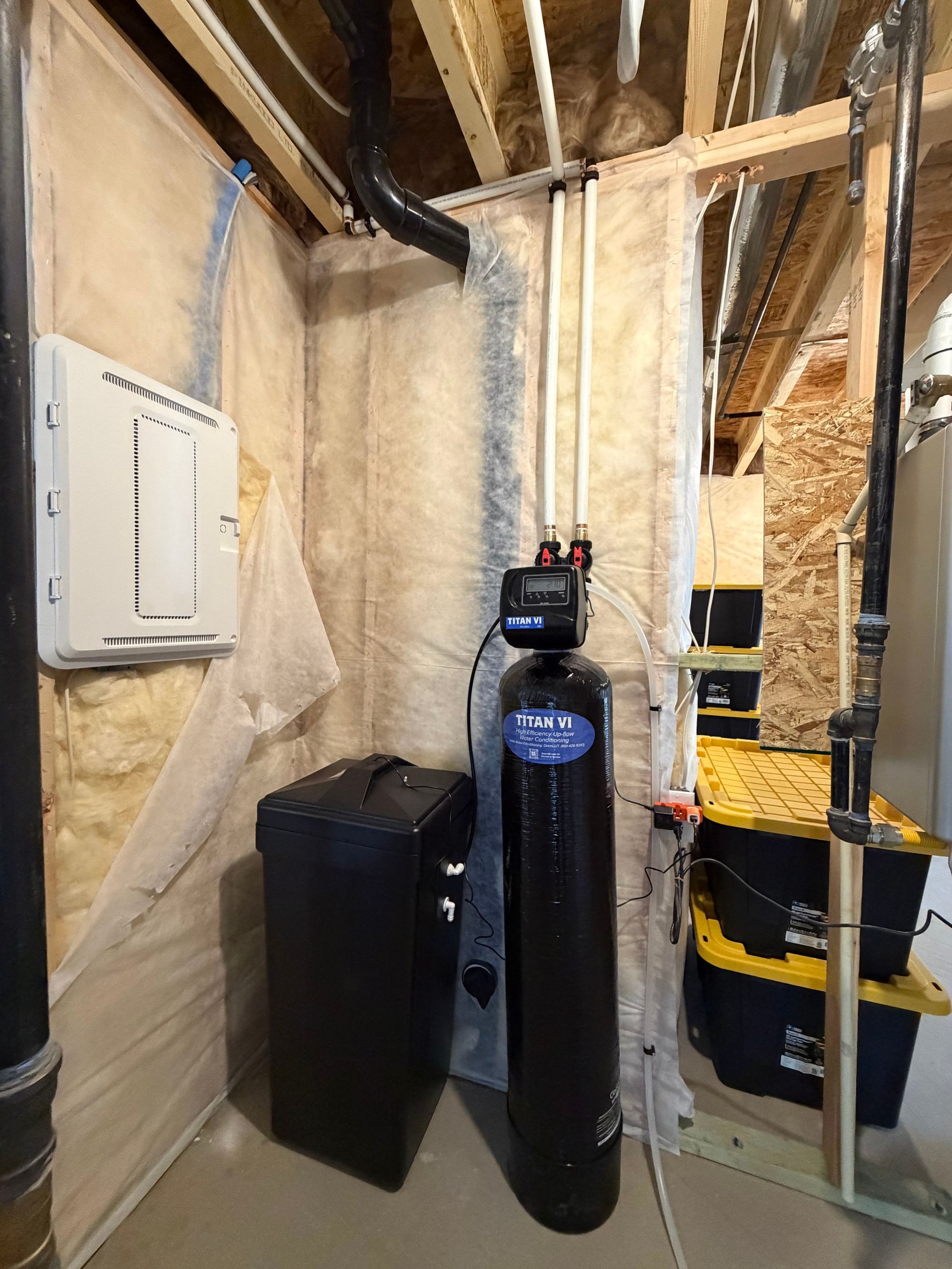 Basement utility area with water softener tank, black bin, and exposed framing beside electrical panel