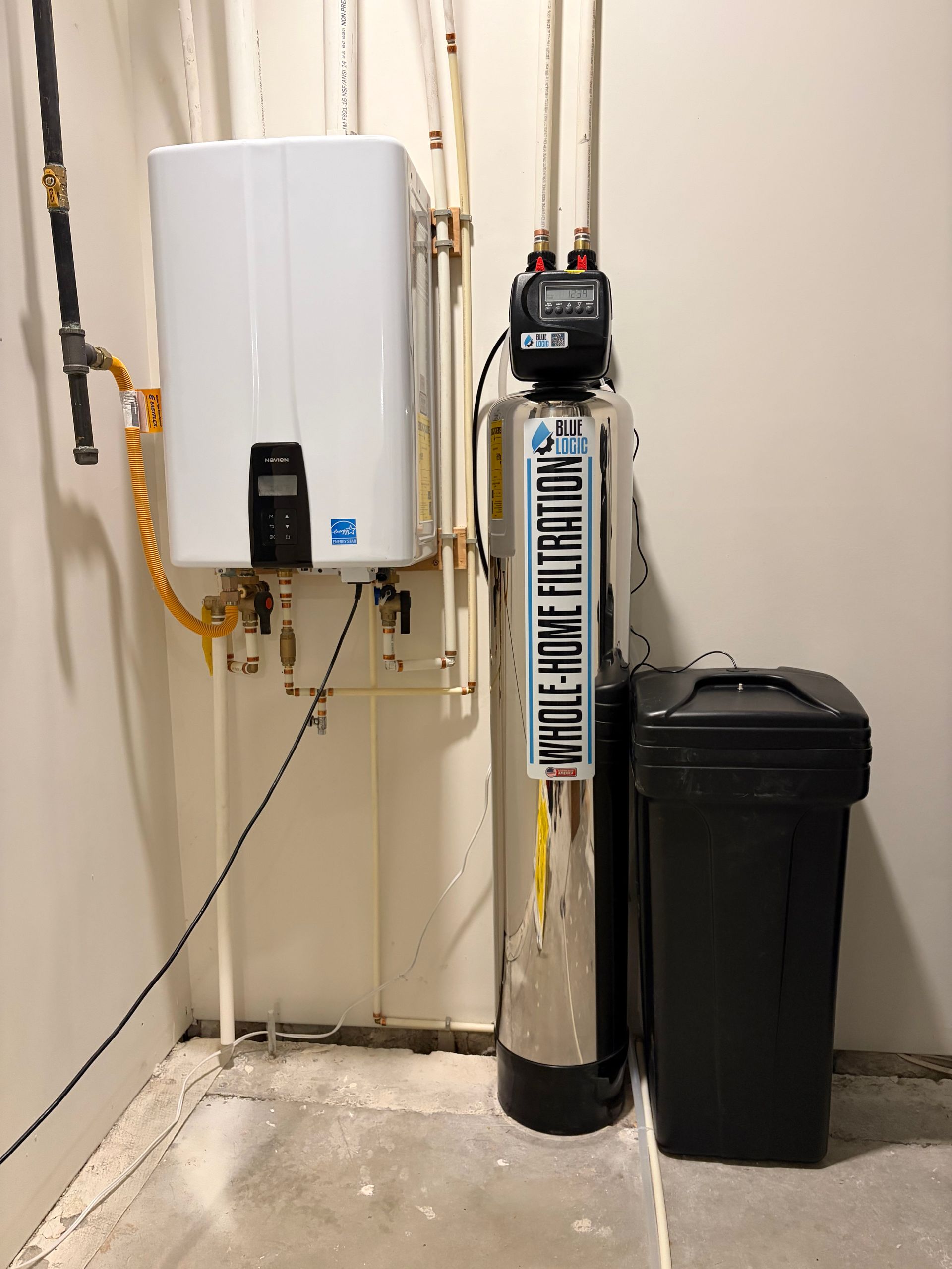 Utility room with a wall-mounted water heater, water softener tank, and trash bin beside plumbing.