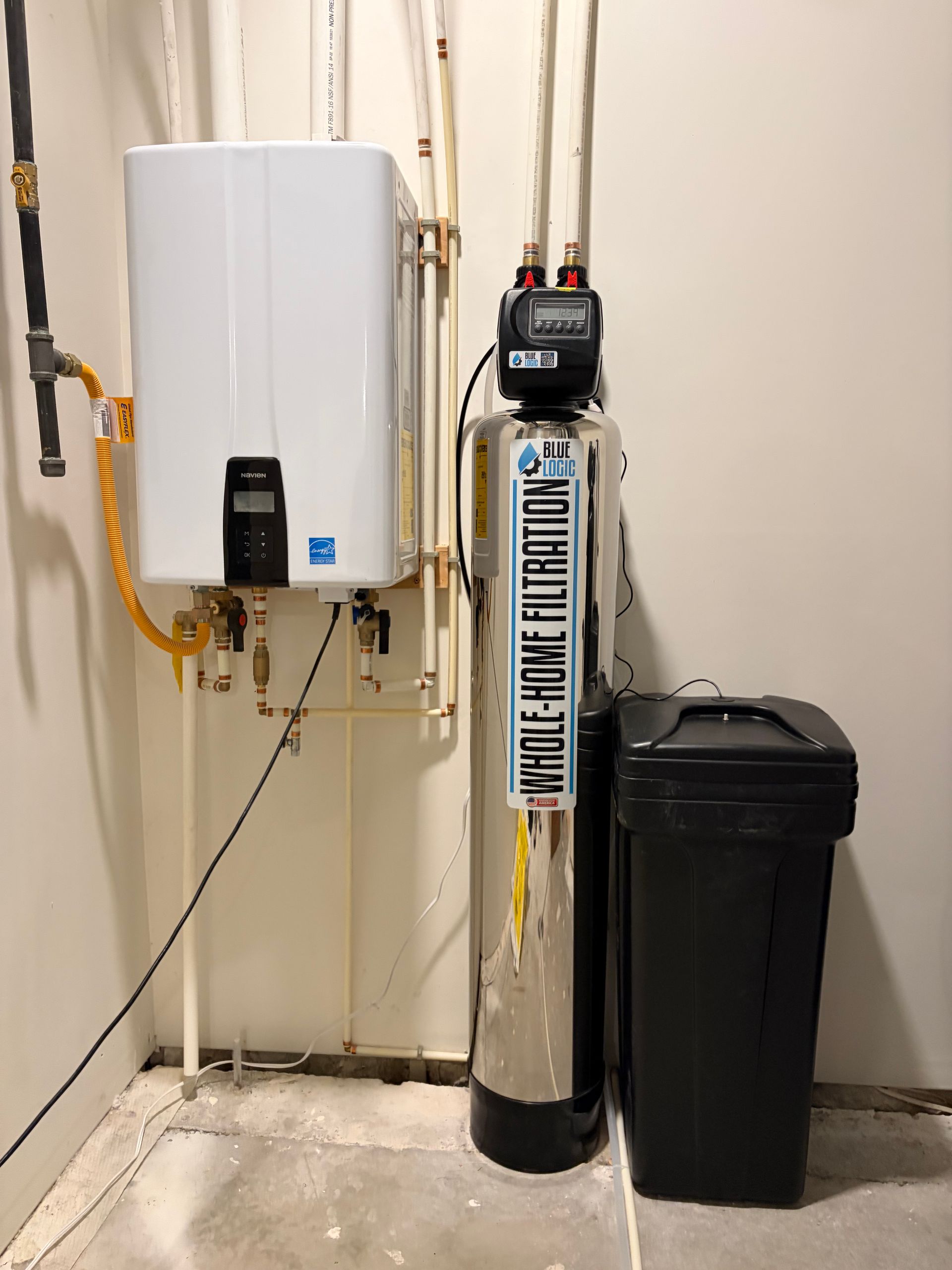 Utility room with wall-mounted water heater, tall cylindrical tank, and a black storage bin against a beige wall