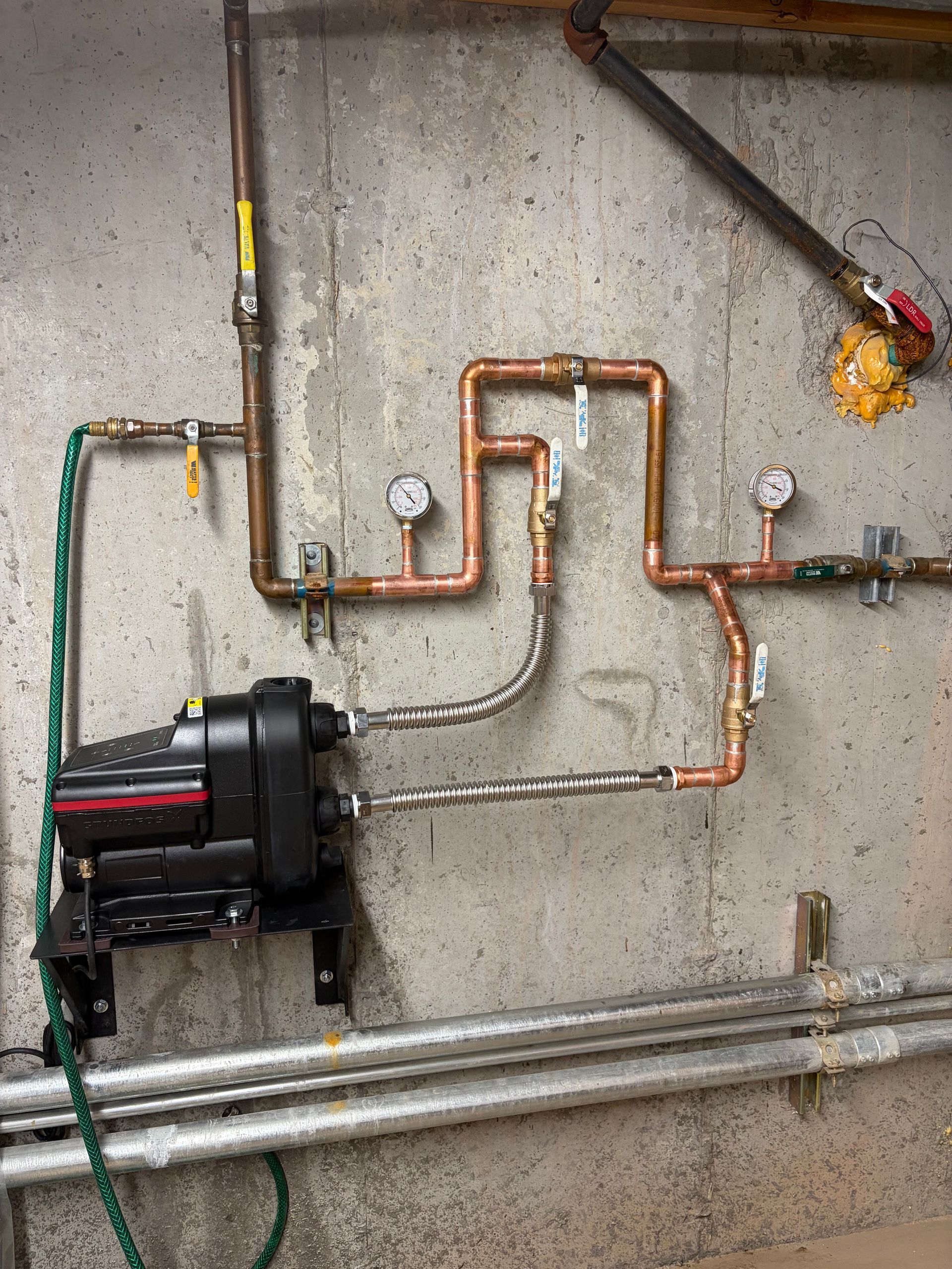 Utility pipes and a black pump mounted on a concrete wall, with copper tubing and gauges.