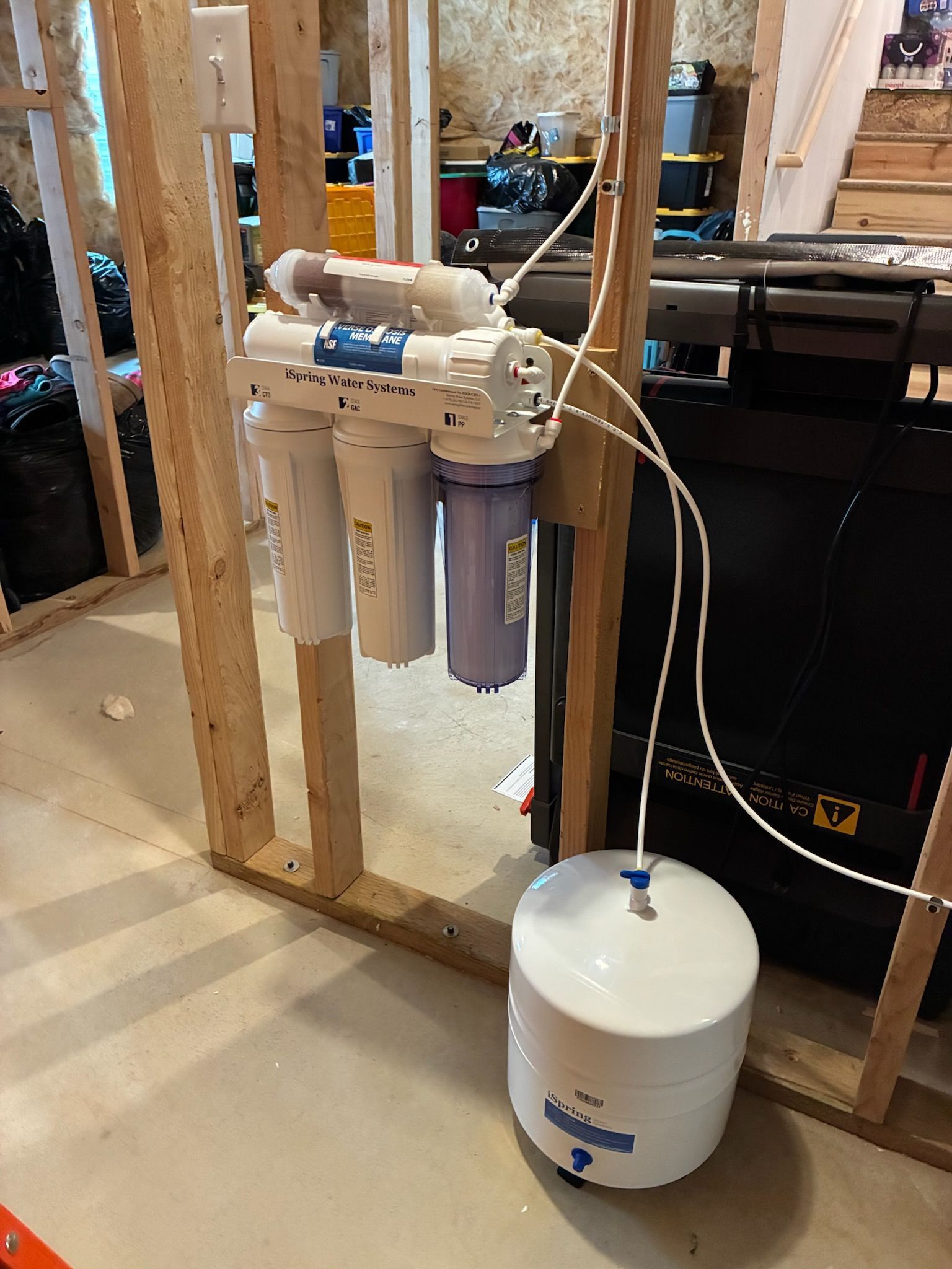 Basement utility setup with water filtration tanks, pipes, and a white pressure tank beside exposed wooden studs