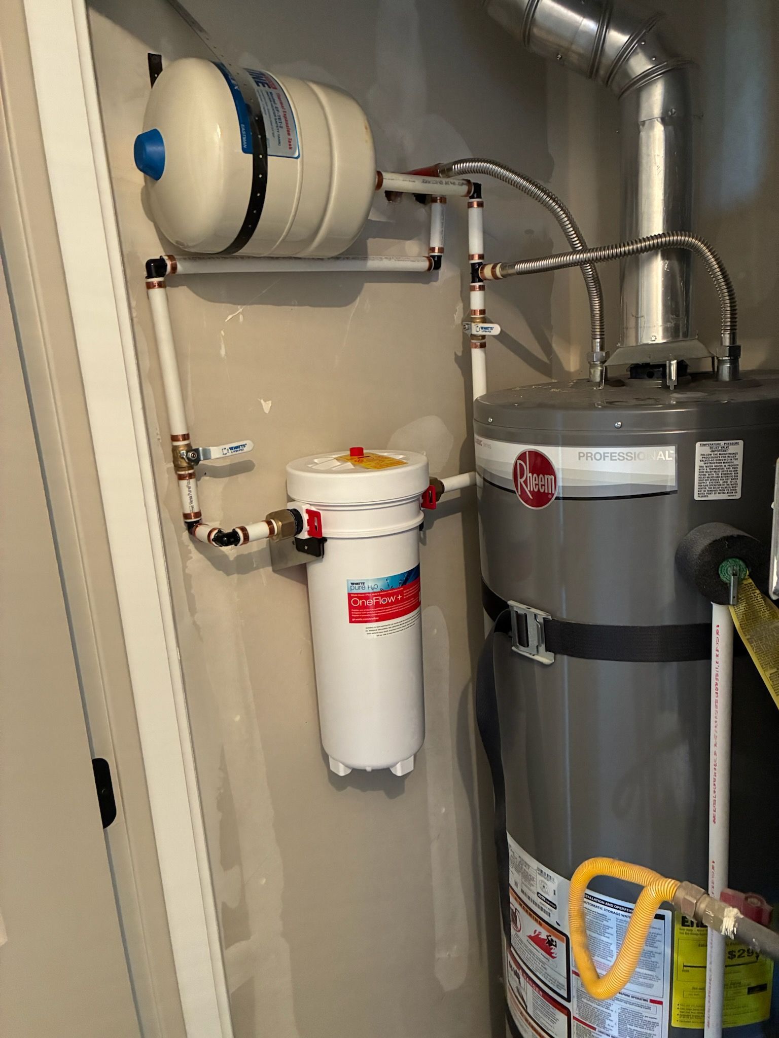 Water treatment equipment in a utility closet, with pipes, a white filter tank, and a gray storage tank.