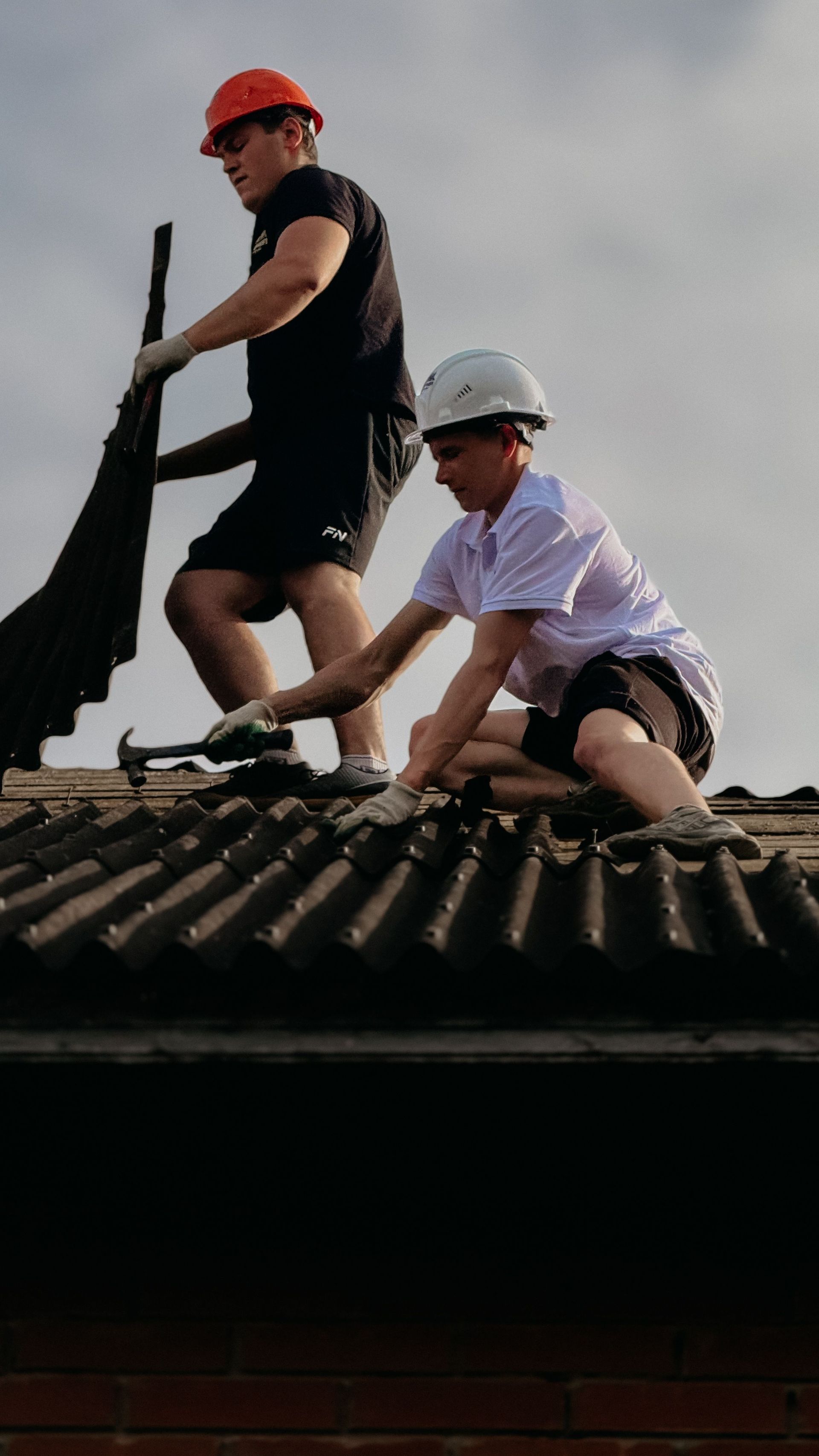 Two workers on a roof wearing hard hats, lifting roof tiles and tools.