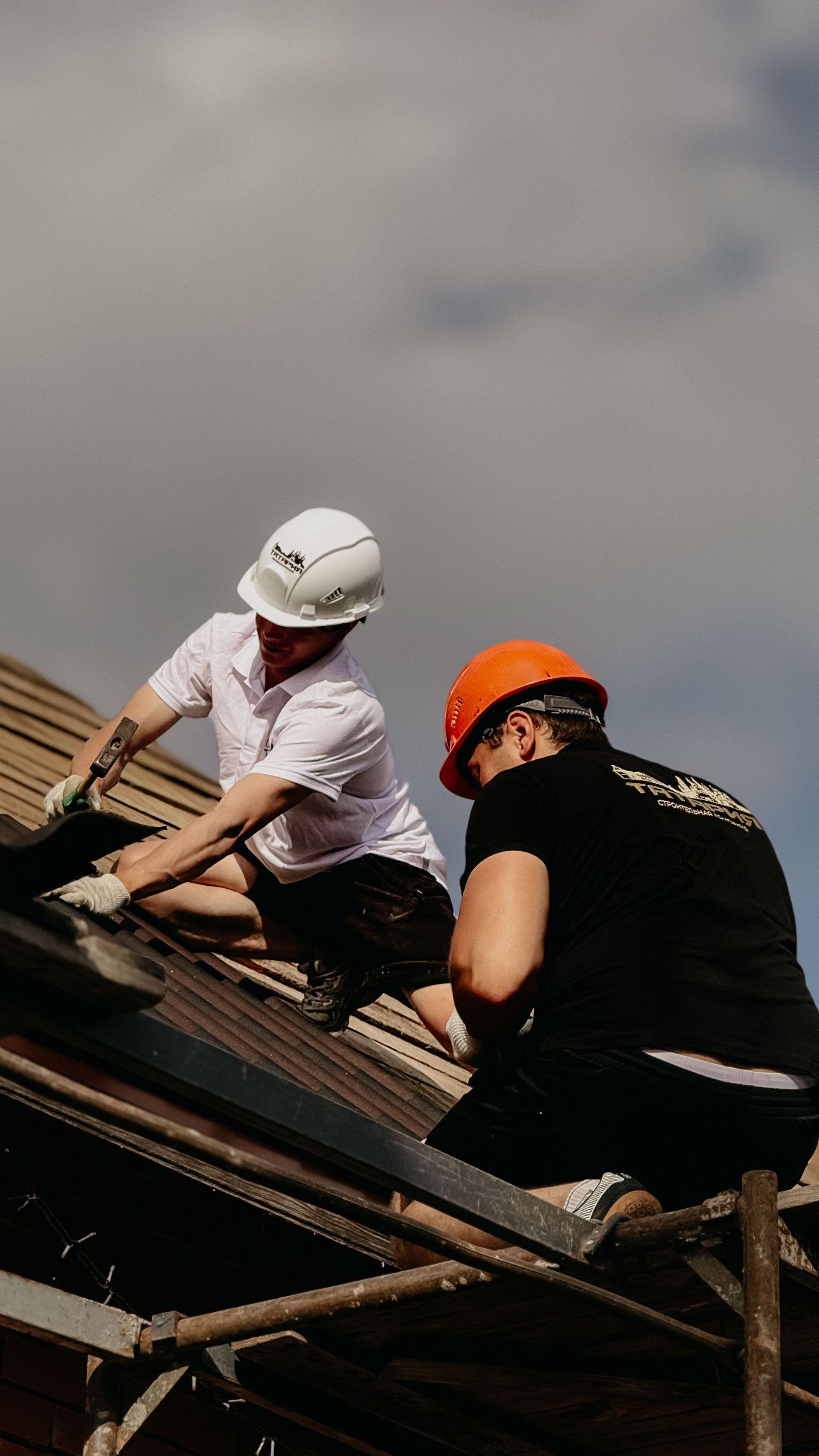 Two workers in white and orange hard hats repair a roof under a cloudy sky.