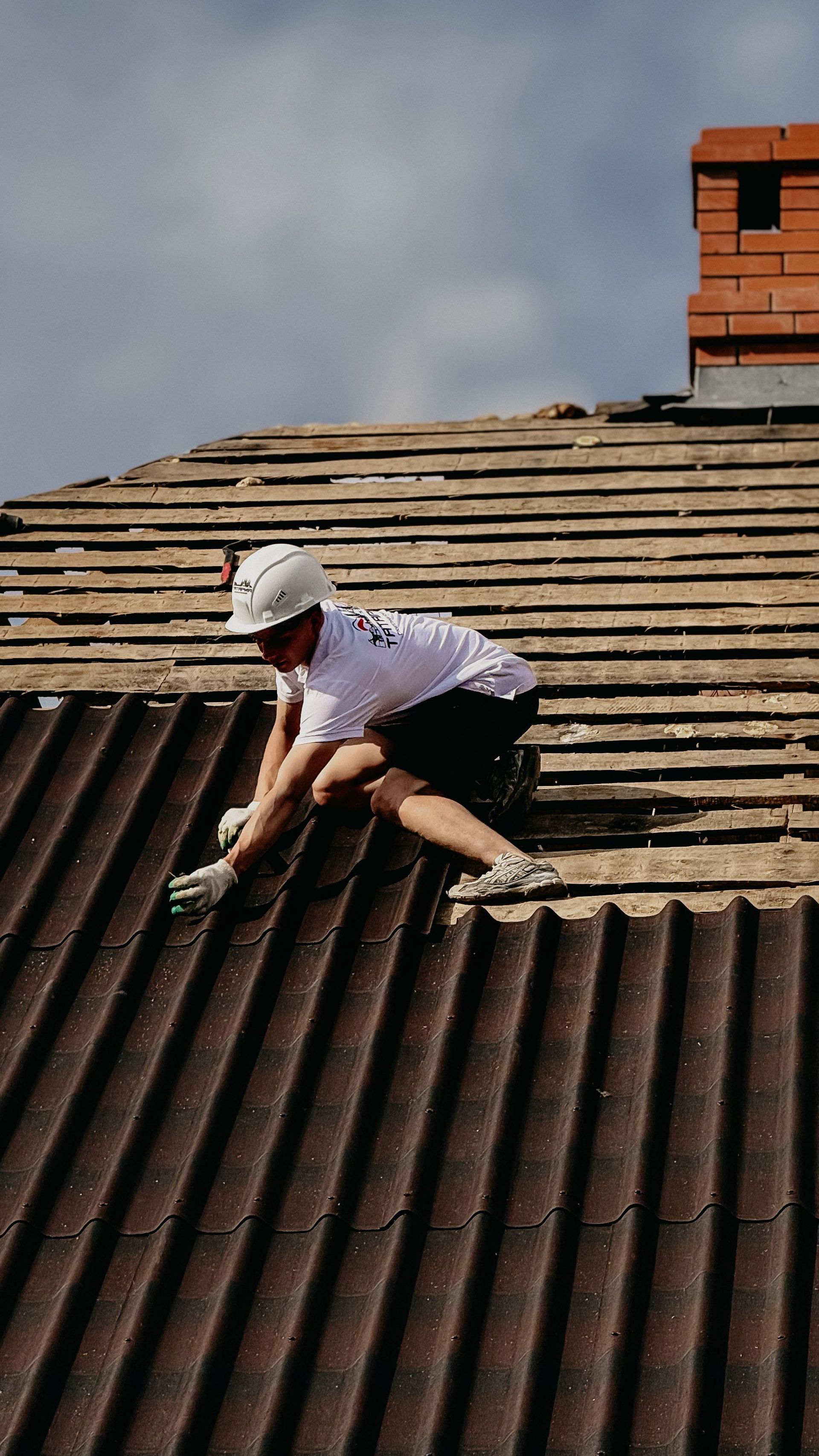 Worker repairing a corrugated roof beside a red brick chimney under a cloudy sky