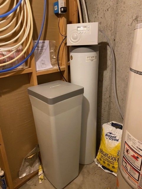 Utility closet with a water softener, trash bin, hoses, and cleaning supplies in a narrow corner.