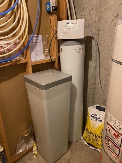 Laundry room corner with water softener tank, trash bin, and coiled hoses on wall shelves.
