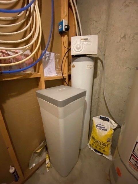 Basement utility area with water softener, pipes, and storage bags against unfinished walls.
