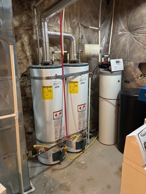 Two water heaters and a water softener in a basement utility room with exposed insulation and piping