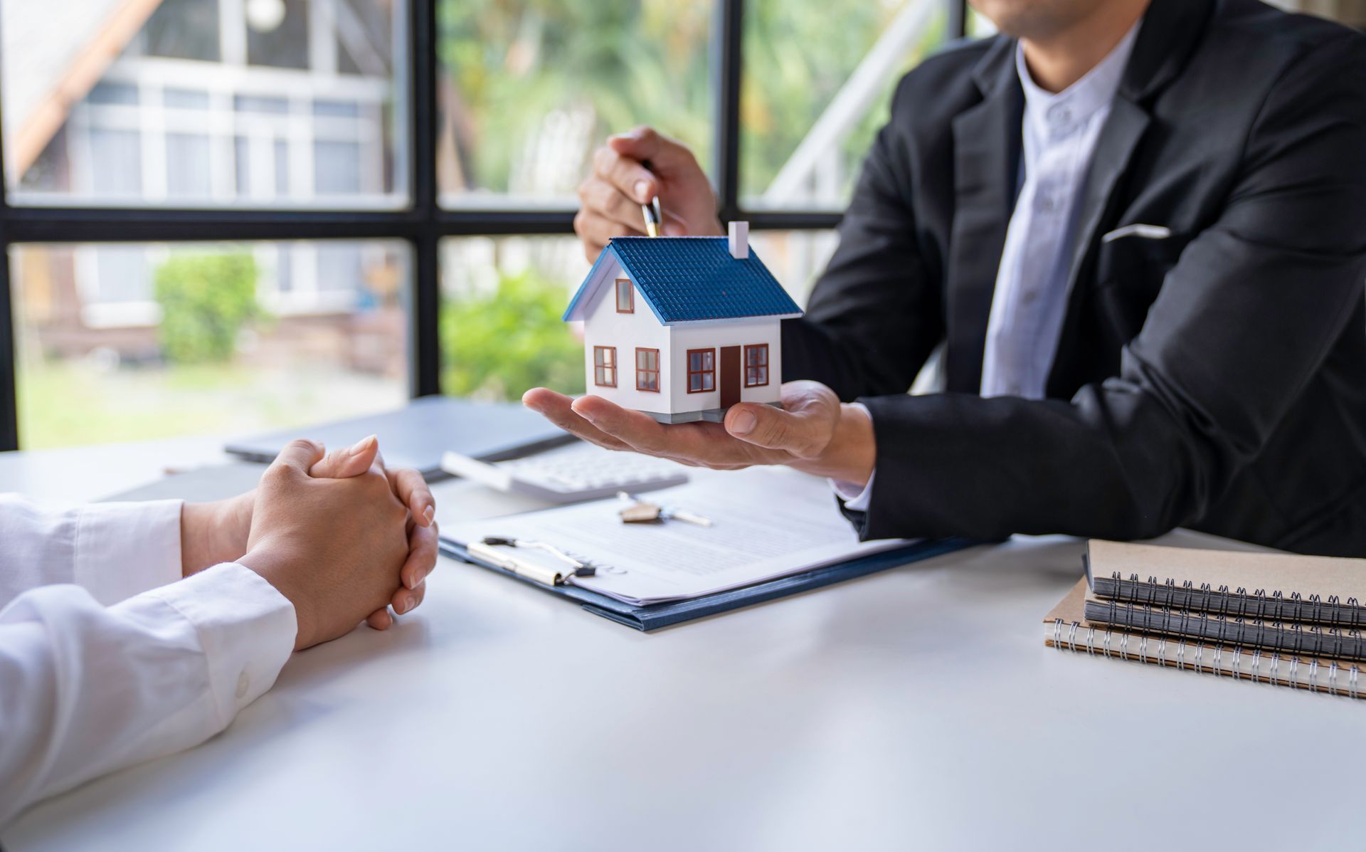 Real estate agent holding house model; client reviews paperwork at a table with a window. Real estate agent holding house model; client reviews paperwork at a table with a window.