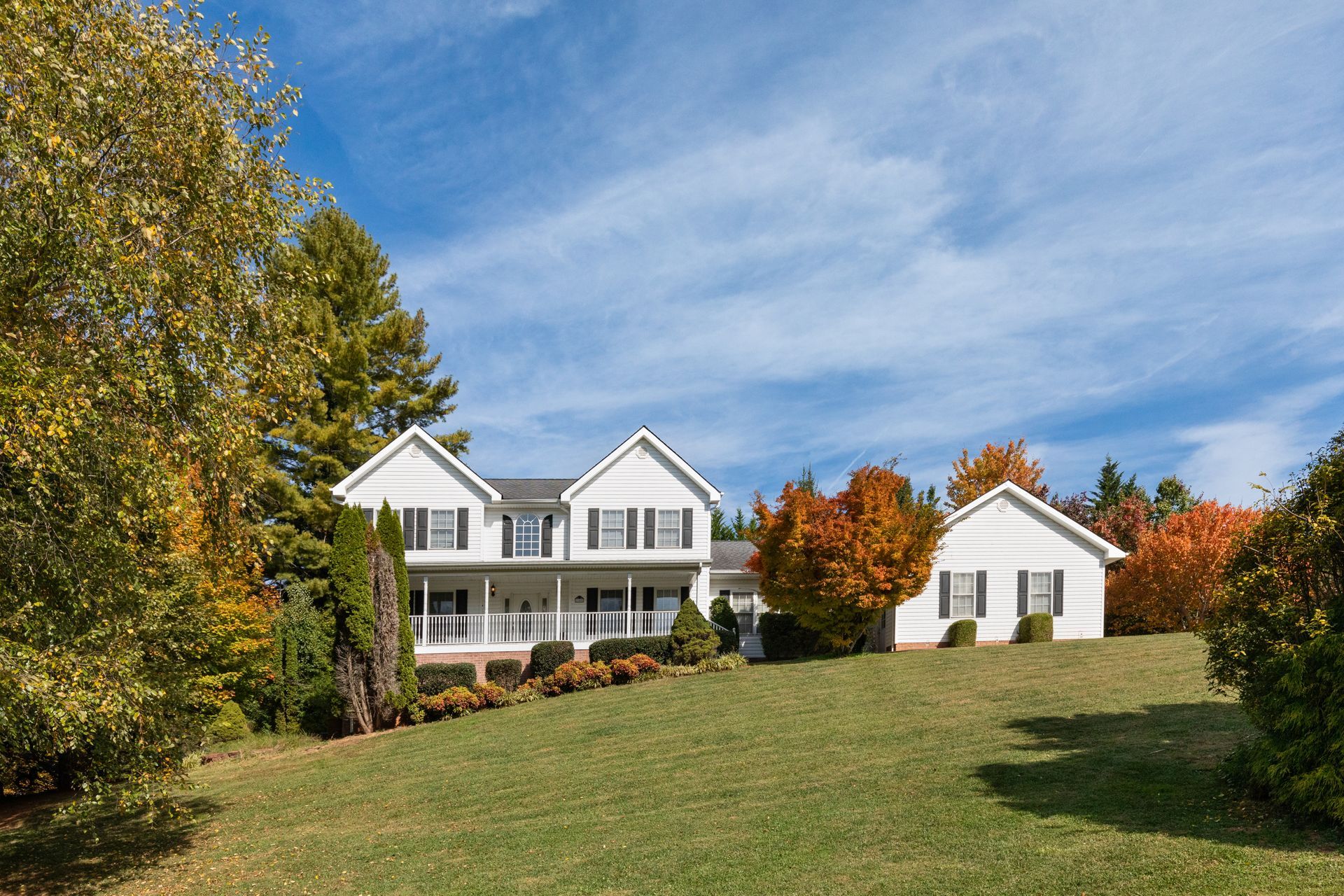 White house with black shutters, on a grassy hill, surrounded by autumn trees, against a blue sky. White house with black shutters, on a grassy hill, surrounded by autumn trees, against a blue sky.