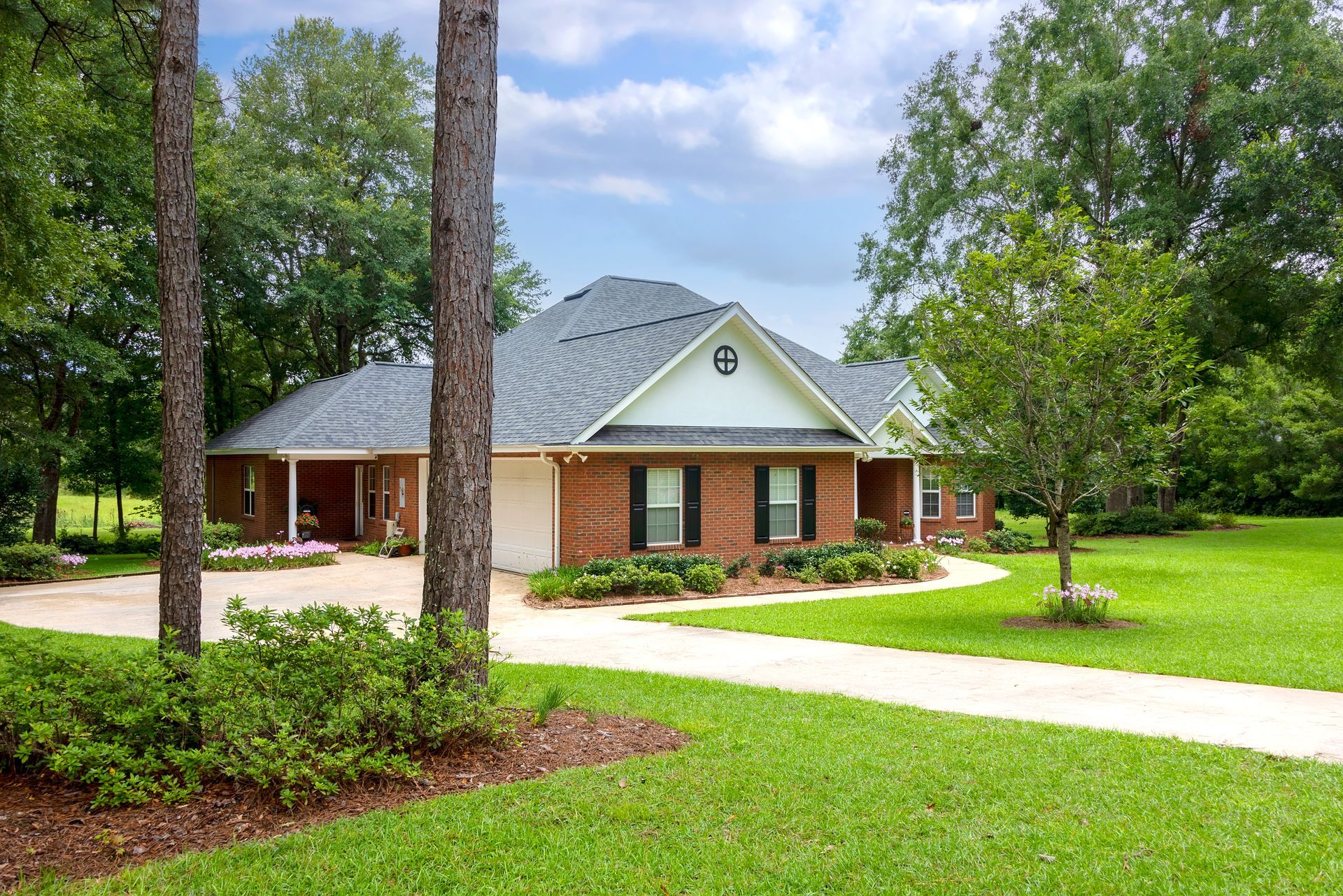 A brick home with a gray roof and circular drive, surrounded by trees and a green lawn under a cloudy sky. A brick home with a gray roof and circular drive, surrounded by trees and a green lawn under a cloudy sky.