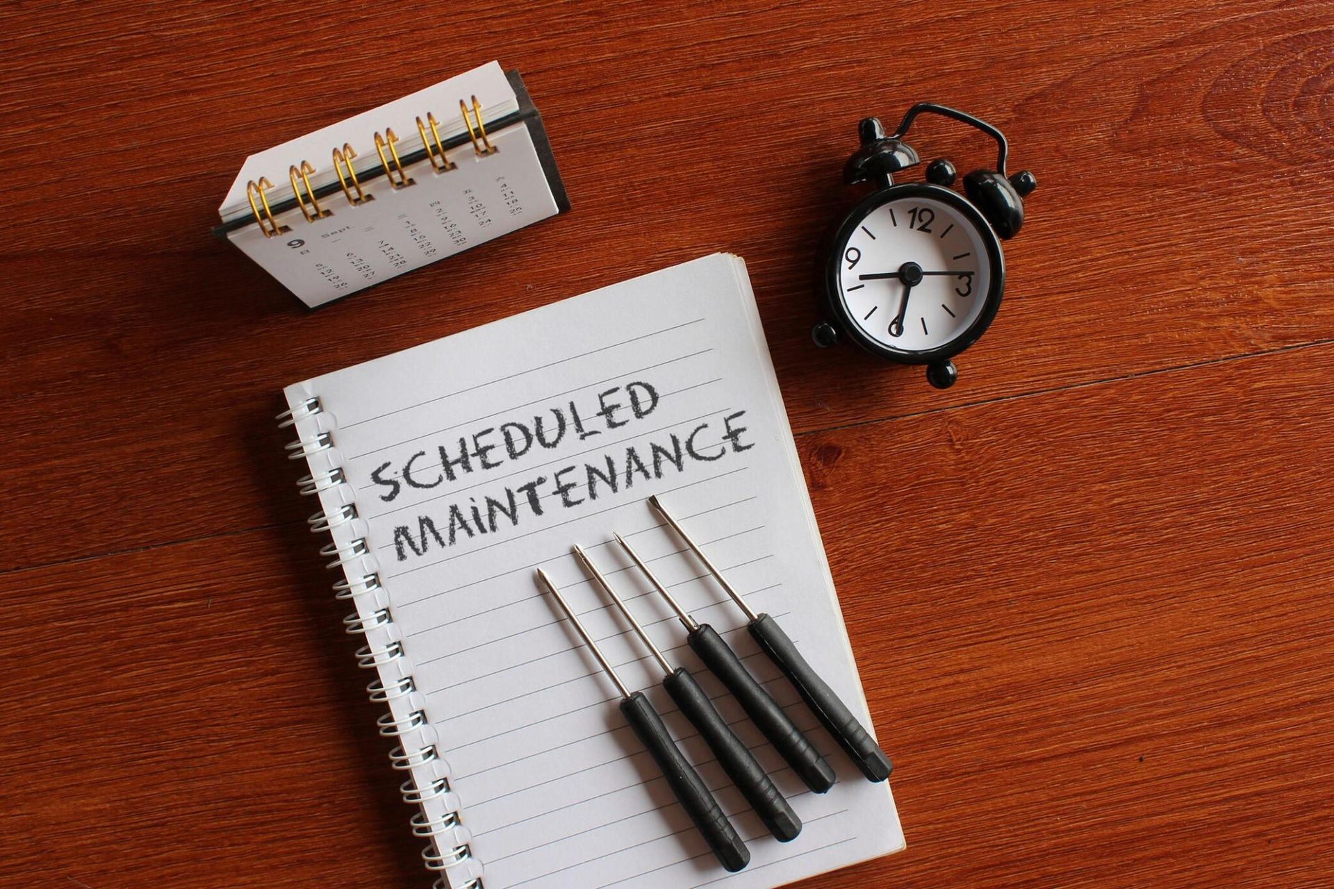 A notebook with scheduled maintenance written on it next to a clock and screwdrivers.