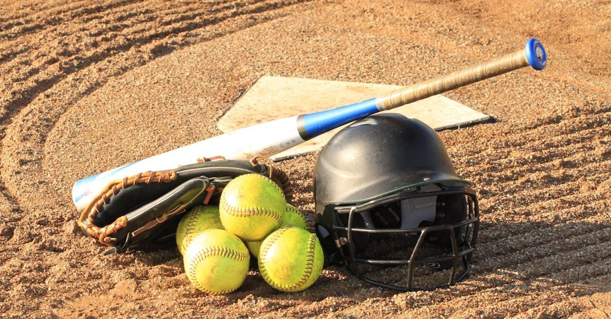 Baseball bat, helmet, glove, and three softballs on a dirt infield