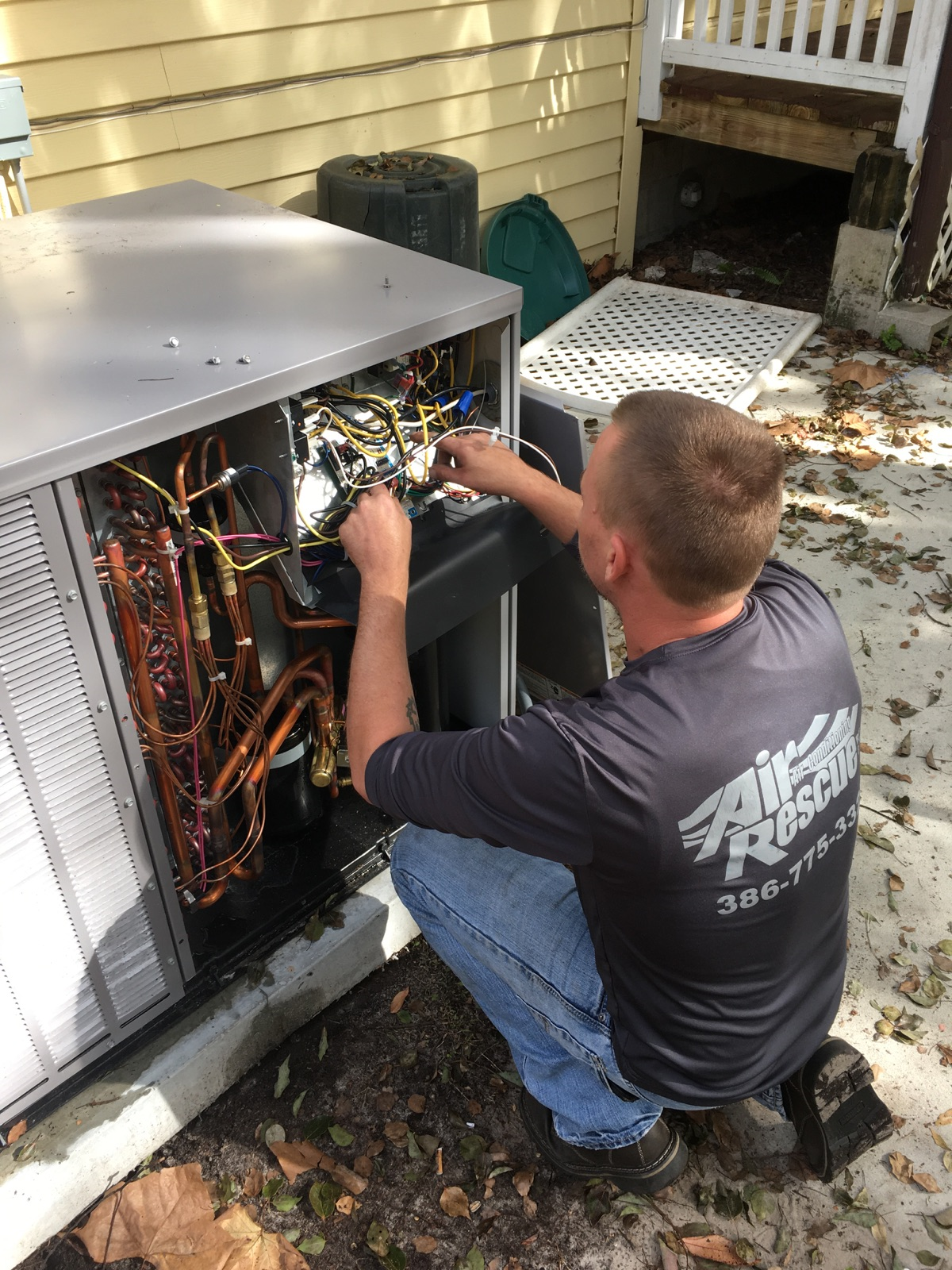 A technician kneels outdoors, repairing the wiring inside the open side panel of a residential air conditioning unit.