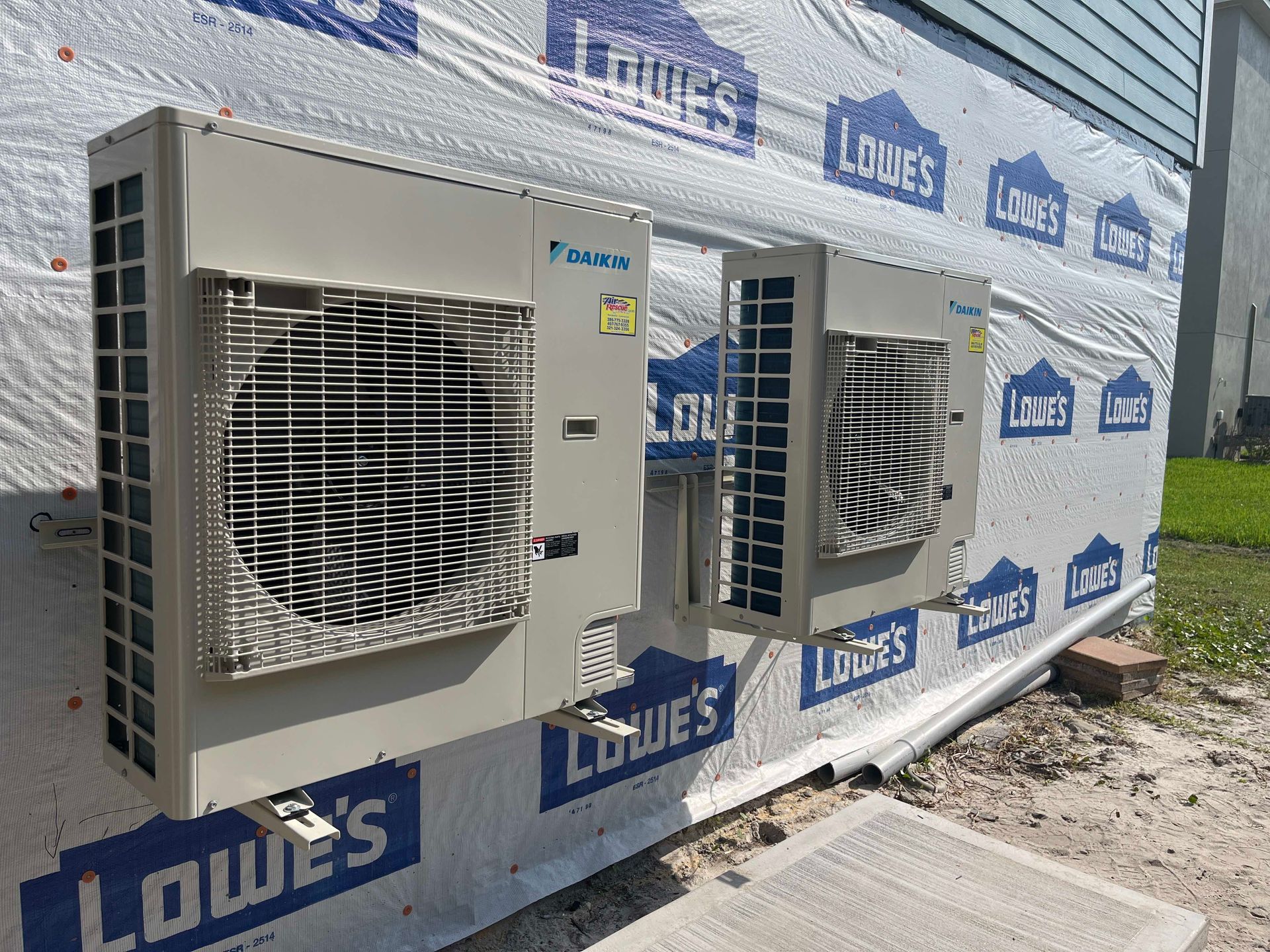 Two beige Daikin air conditioning units mounted to the exterior wall of a building wrapped in Lowe's branded house wrap.