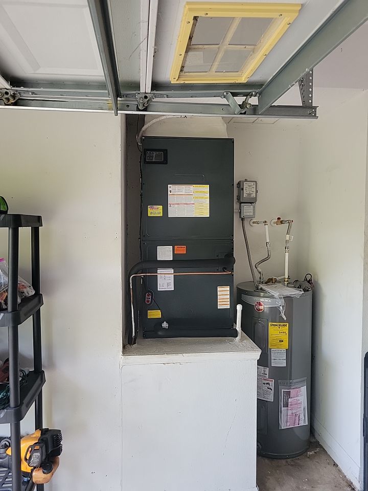 A grey HVAC air handler sits on a white raised platform next to a tall, grey water heater in a residential garage.