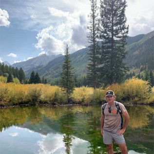 A person wearing a hat and backpack stands by a mountain lake surrounded by golden foliage and tall evergreen trees.