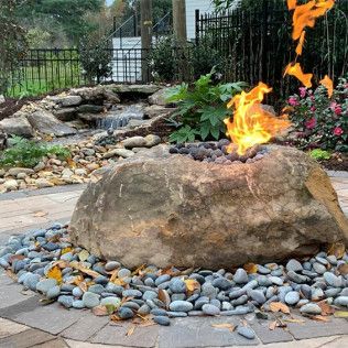 A large natural boulder fire pit with flames burning on top, surrounded by smooth gray stones in a landscaped backyard.
