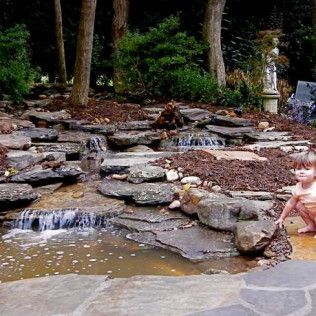 A small child plays by a multi-tiered rock water feature set in a mulched garden with trees and a statue in the background.