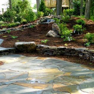 A patio of irregular stone pavers sits before a terraced garden with mulch, rock accents, and a small waterfall.