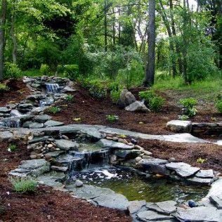 A stone waterfall cascades into a small pond surrounded by mulch and lush green trees in a wooded landscape.