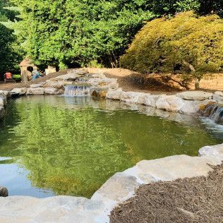 A tranquil pond with cascading stone waterfalls, surrounded by large rocks and lush green trees.