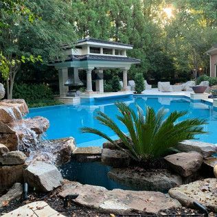 A resort-style pool with a rock waterfall, a central decorative pavilion, and lush landscaping in a backyard setting.