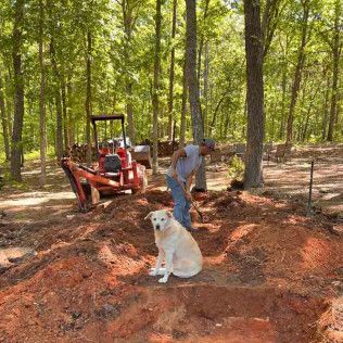 A person uses a shovel near a red backhoe in a wooded area, while a cream-colored dog sits in the foreground on loose dirt.