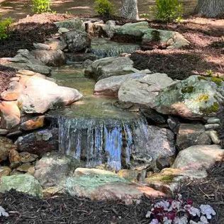 A small man-made waterfall flows over mossy grey rocks into a shallow pool surrounded by dark mulch and greenery.