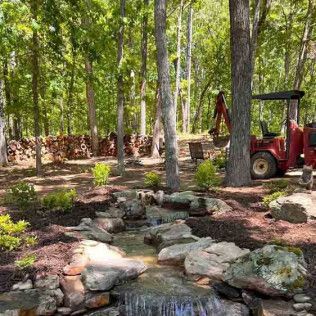 A small waterfall flows into a rock-lined stream in a wooded backyard, with a red utility tractor nearby.