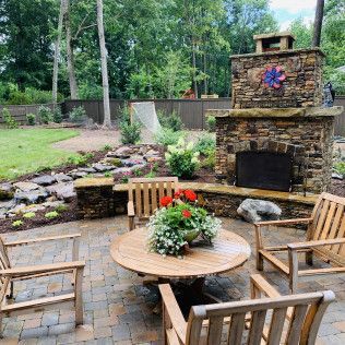 A circular wooden table and chairs sit on a stone patio in front of an outdoor stone fireplace in a landscaped backyard.