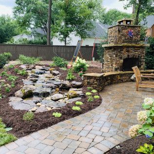A backyard stone fireplace and patio with a small tiered water feature surrounded by mulch and greenery.