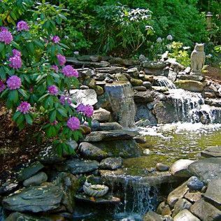 A tiered stone waterfall flows into a pond beside a vibrant bush with purple rhododendron flowers and an owl garden statue.