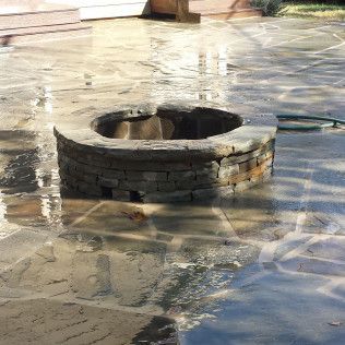 A circular stone fire pit sits on a wet, flagstone patio in a backyard.