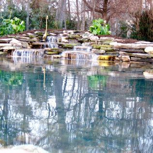 A tiered stone waterfall flows into a clear pond, reflecting the surrounding bare trees and foliage.
