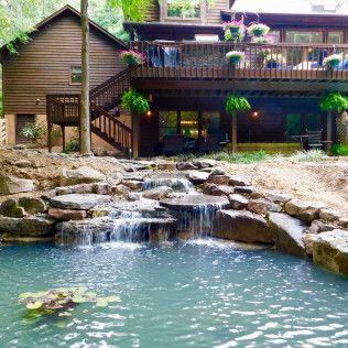 A multi-level wood home with a deck overlooks a tiered stone waterfall cascading into a pond with lily pads.