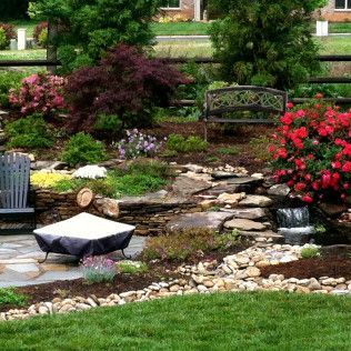 A garden with a stone patio, fire pit, bench, small waterfall, red flowering bushes, and a dark leafy tree.